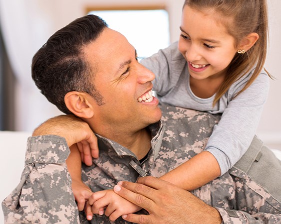 A man in military fatigues smiles at a young girl on his shoulders